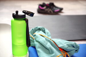 Eine grüne Wasserflasche ist im Vordergrund, daneben drei Gymnastikmatten und ein Handtuch. Im Hintergrund, verschwommen, ist ein Paar Sportschuhe zu erkennen.