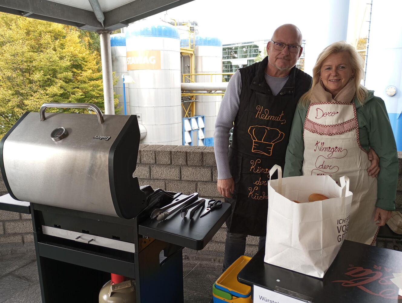 Helmut und Doro stehen neben einem Grill, mit einer großen Brötchentüte. Beide tragen eine Schürze mit ihren Namen und der Aufschrift "König/Königin der Küche".