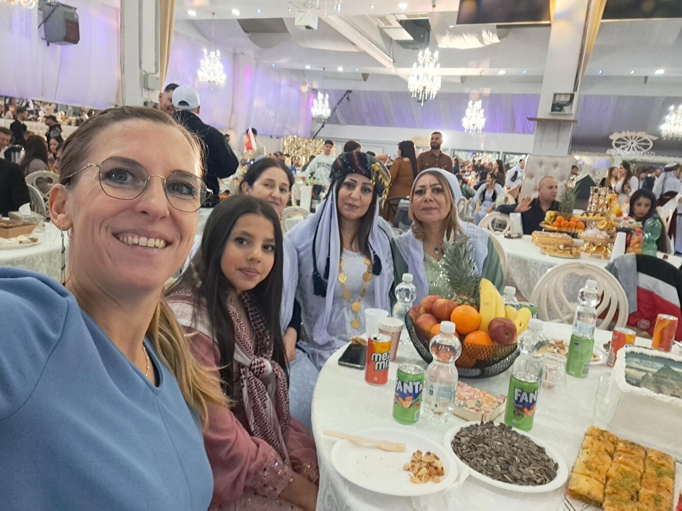 Eine große Festhalle. Ann Jerusalem im Vordergrund, mit vier jesidischen Frauen in traditioneller Kleidung hinter ihr. Alle sitzen an einem Tisch mit Getränken, Obstschale und verschiedenem anderen Essen.