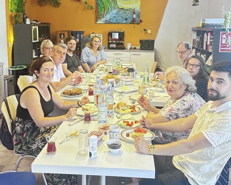 Gemeinsames Frühstück in einem Gemeinschaftsraum: Mehrere erwachsene Personen sitzen an einem großen Tisch mit Speisen und Getränken.