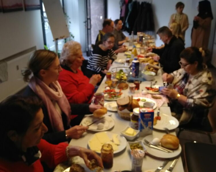 Gemeinsames Frühstück in einem Gemeinschaftsraum: Mehrere erwachsene Personen sitzen an einem großen Tisch mit Speisen und Getränken.