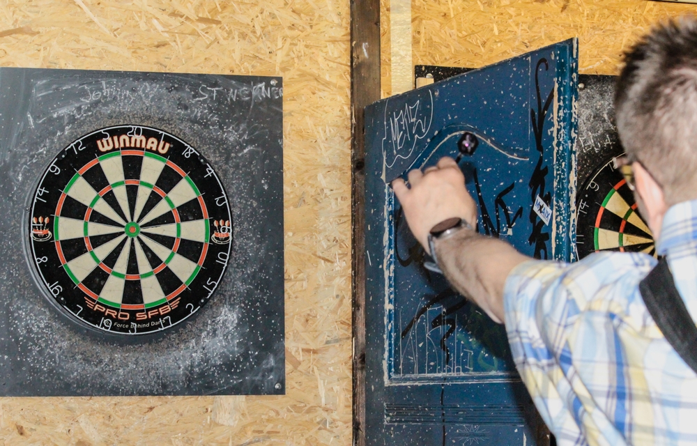 Eine Person steht vor einer an der Wand befestigten Dartscheibe und hält einen Darts-Pfeil in Wurfhaltung